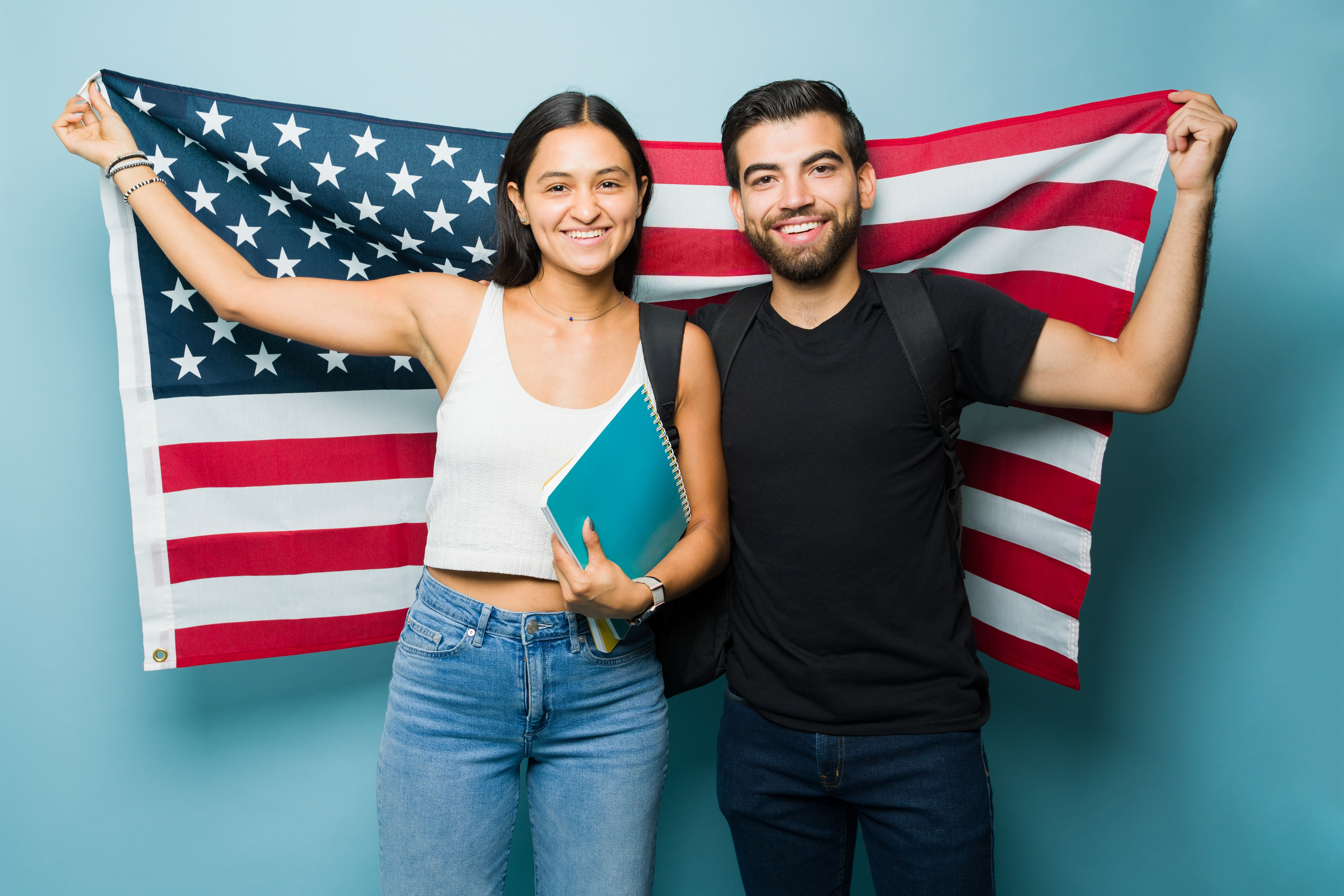 Two young adults smiling and holding an American flag, one carrying a notebook and backpack, representing immigration success, citizenship, and achieving the American dream.